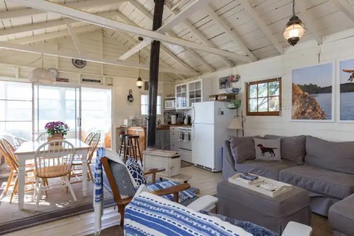 Cozy cabin interior with a living area, dining table, and kitchen, featuring light wood accents and bright natural light.