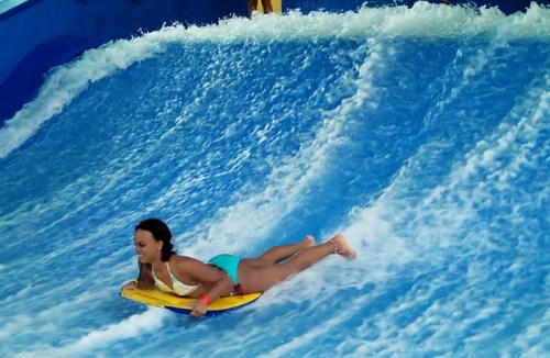 A person rides a bodyboard on a wave simulator, surrounded by splashing water.