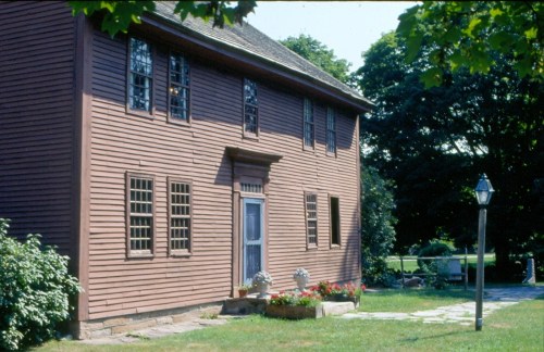 A historic brown wooden house with multiple windows, surrounded by greenery and a stone pathway.