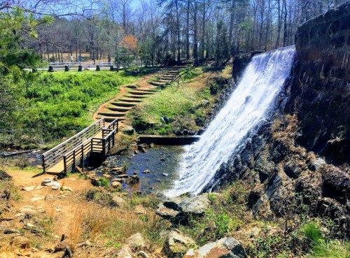 A serene waterfall cascades over rocks, surrounded by greenery and a wooden bridge leading to a path.