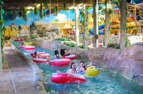 People float on colorful inner tubes in a lazy river surrounded by tropical decor and water slides.
