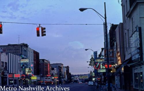 A vintage street scene featuring neon signs and storefronts at dusk, with traffic lights and a clear sky.