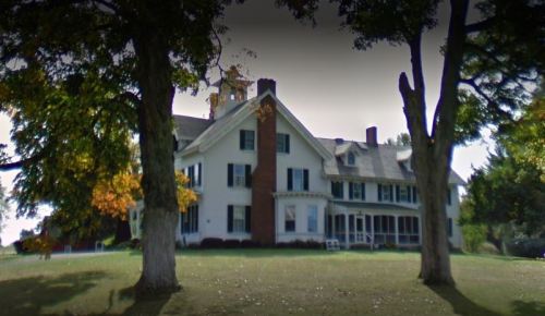 A large, historic white house with green shutters, surrounded by trees and a grassy lawn.