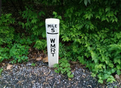 A white mile marker post labeled "MILE 5 WMRT" surrounded by green foliage and gravel.