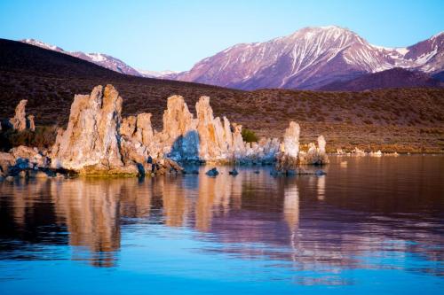 Rock formations rise from a calm lake, reflecting mountains with snow-capped peaks in the background during sunset.