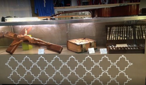 Display case featuring a wooden crucifix, an old book, and a set of silver spoons.