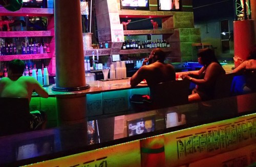 A vibrant bar scene with patrons sitting at the counter, colorful lights, and shelves stocked with drinks.