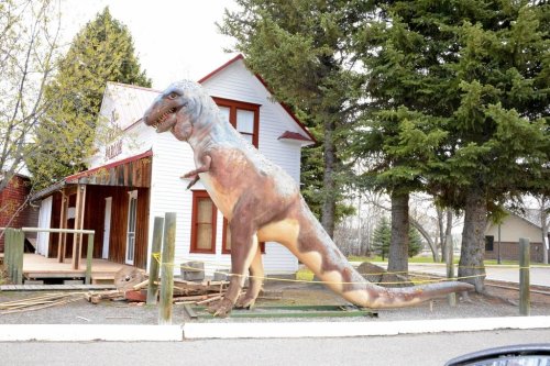 A large dinosaur statue stands outside a wooden building surrounded by trees.