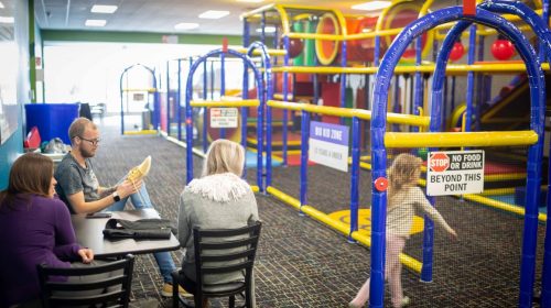 A colorful indoor play area with children’s equipment, while adults sit at a table nearby.