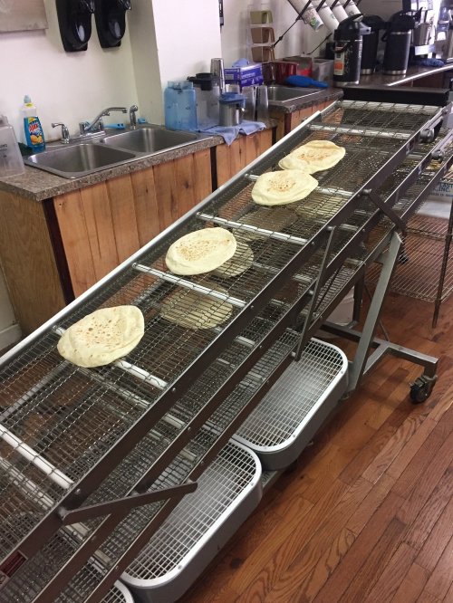 Flatbreads resting on a cooling rack in a kitchen, with sinks and kitchen equipment in the background.