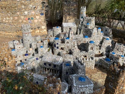 A miniature stone village with blue domes, featuring intricate buildings and pathways, surrounded by greenery.
