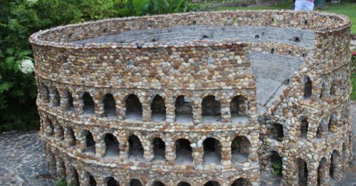 A miniature replica of the Colosseum made from stones, featuring arches and a circular design, set in a garden.