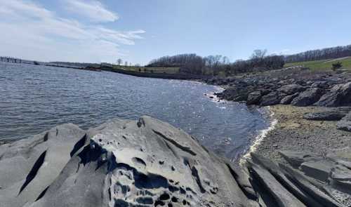 A rocky shoreline with calm water, under a clear blue sky and distant green hills.