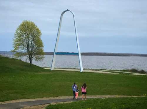 Two people walk along a path near a large white arch structure by a lake, with a tree and cloudy sky in the background.