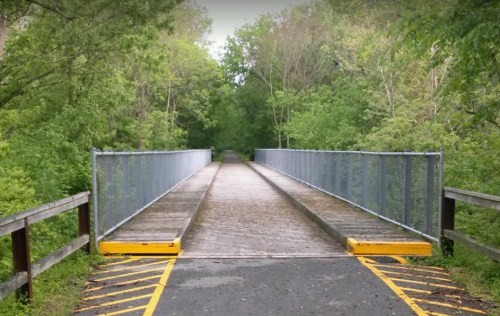 A wooden bridge with metal railings spans a green, tree-lined path, leading into a lush forest.