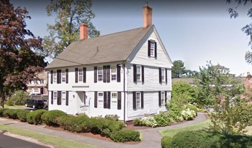 A two-story white house with black shutters, surrounded by greenery and a paved driveway.