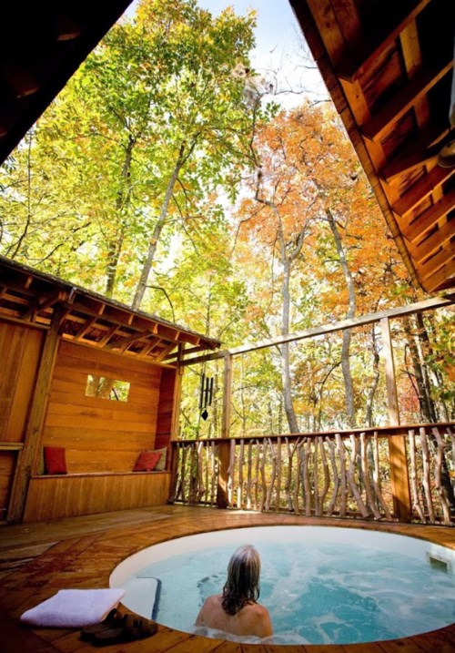A person relaxes in a hot tub surrounded by autumn trees, with wooden walls and a cozy atmosphere.