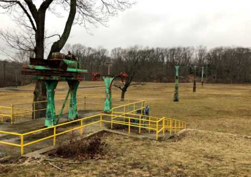 Abandoned amusement park with rusted structures and yellow railings in a grassy field under a cloudy sky.