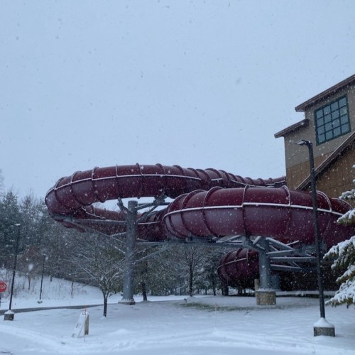 A large, red water slide structure covered in snow, with falling snowflakes and a building in the background.