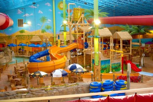 A colorful indoor water park featuring slides, play structures, and seating areas under a bright blue ceiling.