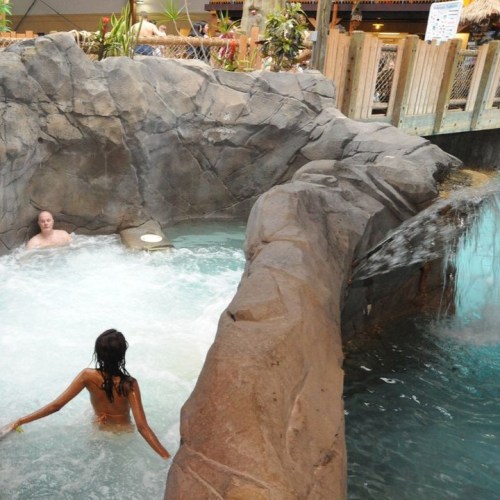 A woman in a swimsuit relaxes in a rocky hot tub, while a man sits nearby in a water-filled area.