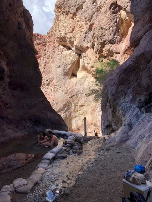 A narrow canyon with colorful rock walls, featuring a small group of people relaxing by a water source.