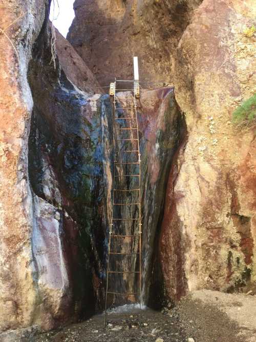 A metal ladder leans against a colorful rock face with water flowing down, surrounded by rugged terrain.