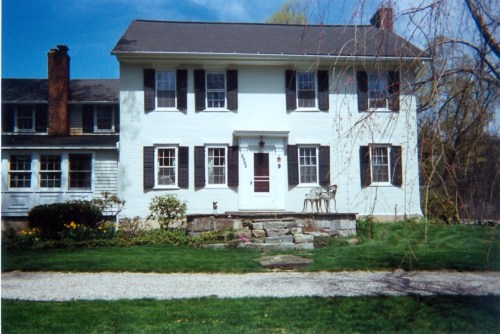 A two-story white house with black shutters, a red door, and a stone pathway surrounded by green grass and flowers.
