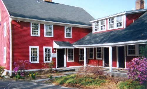 A red two-story house with white trim, featuring a porch and surrounded by blooming flowers.