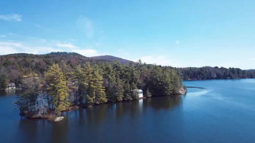 A serene lake surrounded by lush trees and a small island with a white structure, under a clear blue sky.