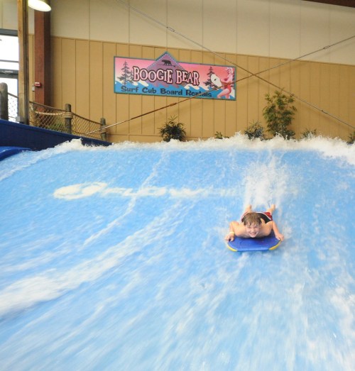 A child rides a boogie board on a wave simulator, splashing water in a fun indoor surf environment.