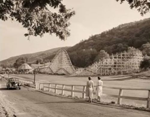 Two people walk along a road near a vintage amusement park with a wooden roller coaster in the background.