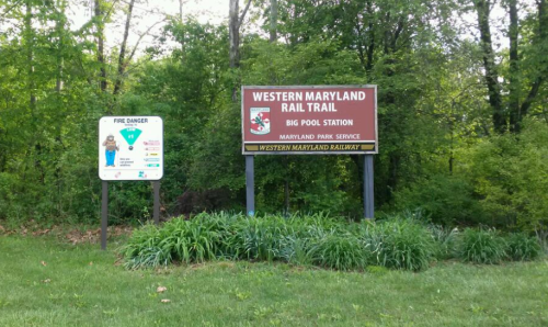 Sign for Western Maryland Rail Trail at Big Pool Station, surrounded by greenery and a fire danger warning sign.