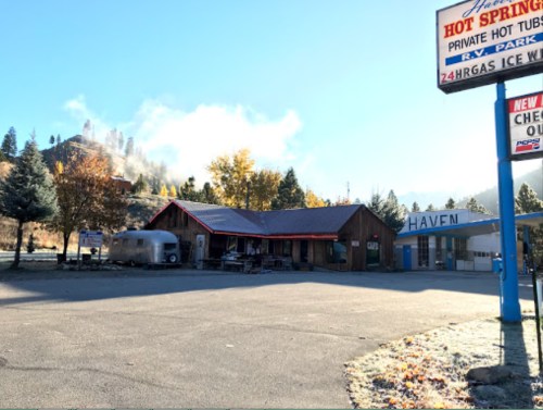 A rustic building with a sign for hot springs, surrounded by trees and mountains, under a clear blue sky.