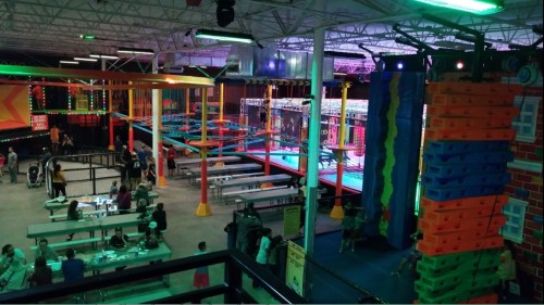 A vibrant indoor trampoline park with colorful equipment, people enjoying activities, and seating areas visible.