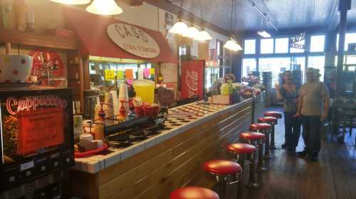 A vintage-style diner with a wooden counter, red stools, and a menu board, featuring customers chatting in the background.