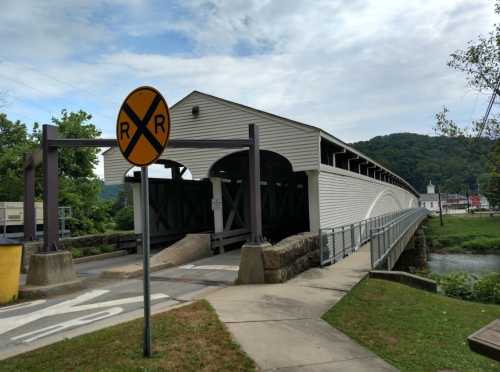 A covered bridge with a railroad crossing sign, surrounded by greenery and a river, under a cloudy sky.