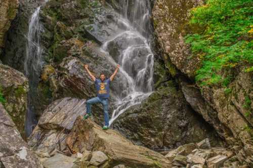 A person stands triumphantly on a rock near a waterfall, surrounded by lush greenery and rugged stone formations.