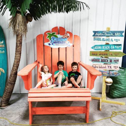 Three children sit on a large orange chair, surrounded by tropical decor, smiling at the camera.