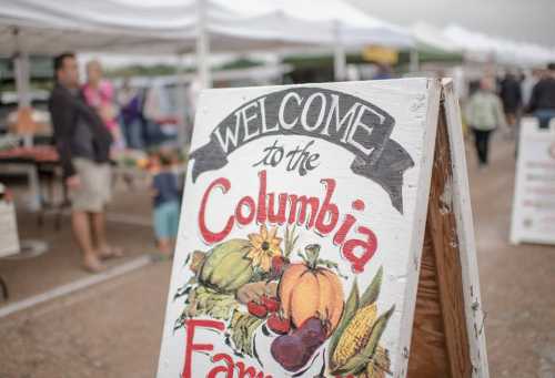 A wooden sign reading "Welcome to the Columbia Farm" with colorful fruit and vegetable illustrations at a farmers market.