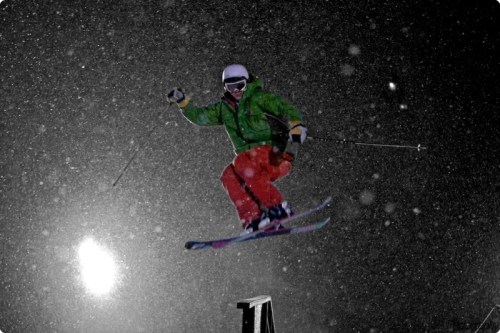 A skier in a green jacket and red pants jumps mid-air during a snowstorm, with snowflakes swirling around.