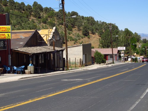A quiet street scene with rustic buildings, trees on a hillside, and a clear blue sky.
