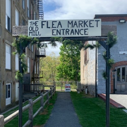 Sign for "The Old Mill Flea Market Entrance" with a pathway leading to the market, surrounded by greenery.