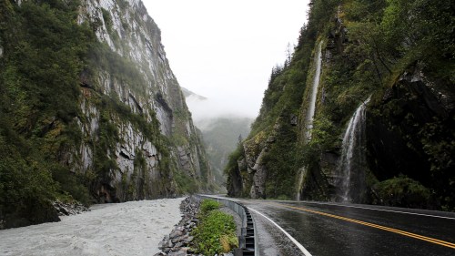 A winding road alongside a river, flanked by steep cliffs and waterfalls in a misty, green landscape.