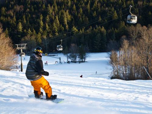 A snowboarder descends a snowy slope with ski lifts and trees in the background on a sunny day.