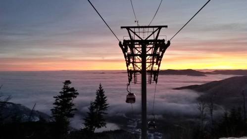 A ski lift tower silhouetted against a colorful sunset over a sea of clouds and distant mountains.