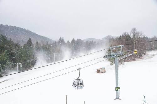 A snowy landscape with a ski lift, snowmaking machines, and mountains in the background under a cloudy sky.
