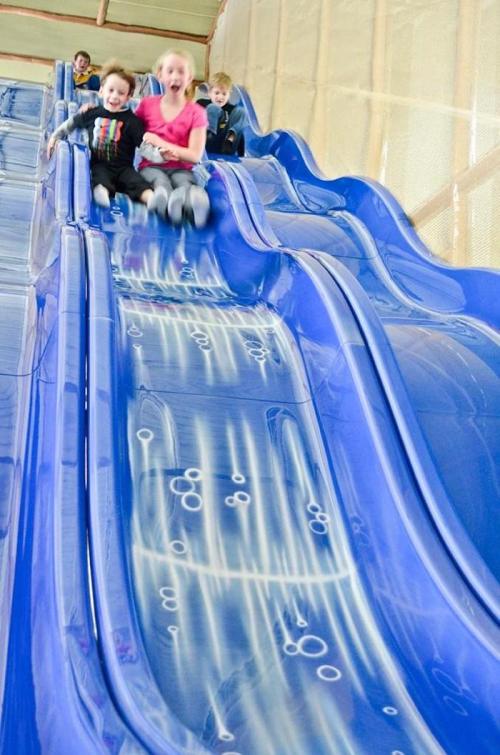 Two children joyfully slide down a blue water slide, with others in the background enjoying the fun.