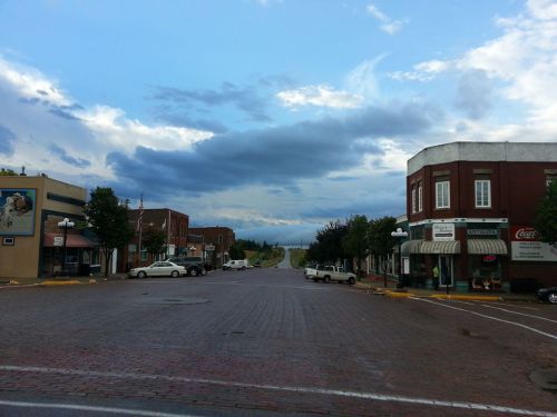 A quiet town square with brick pavement, lined by small shops and under a cloudy sky.