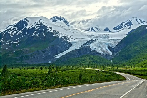 A winding road leads through lush green fields towards snow-capped mountains and a glacier under a cloudy sky.